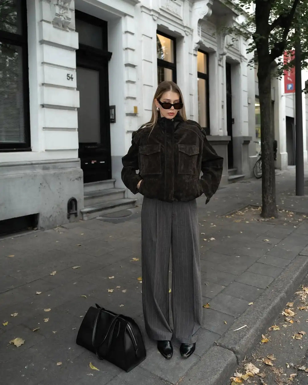 Woman in a dark jacket and gray skirt standing on a city street with a black handbag.