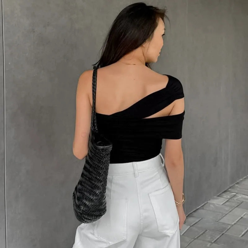 Woman wearing a black one-shoulder top and white pants against a gray wall.
