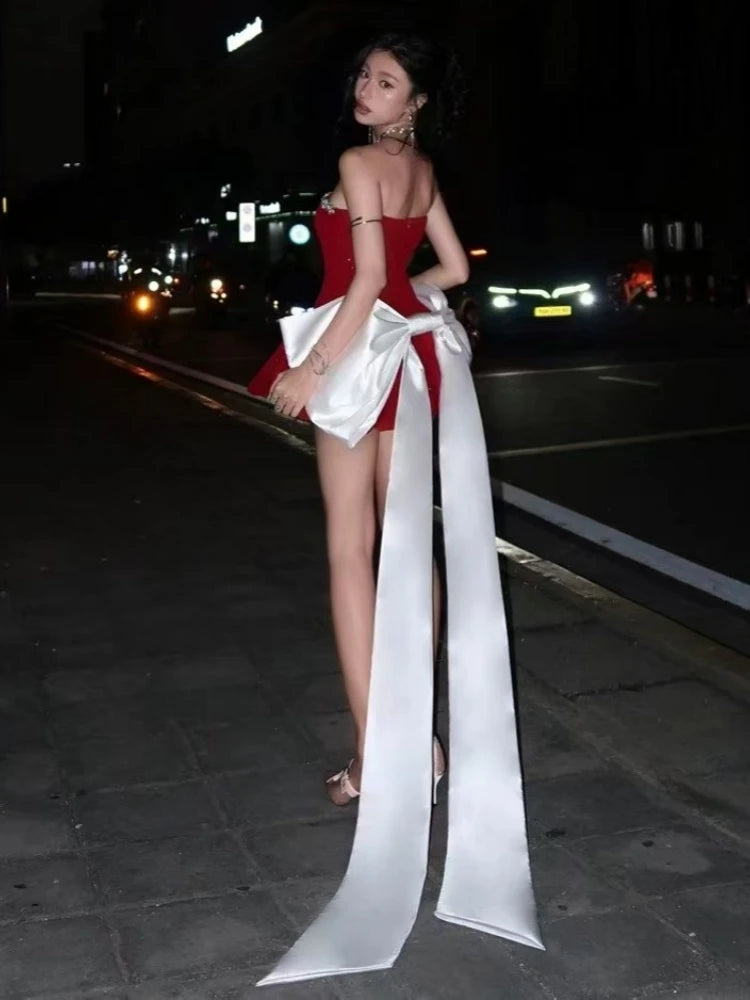 Woman in a red dress with a long white bow on a dark street at night.