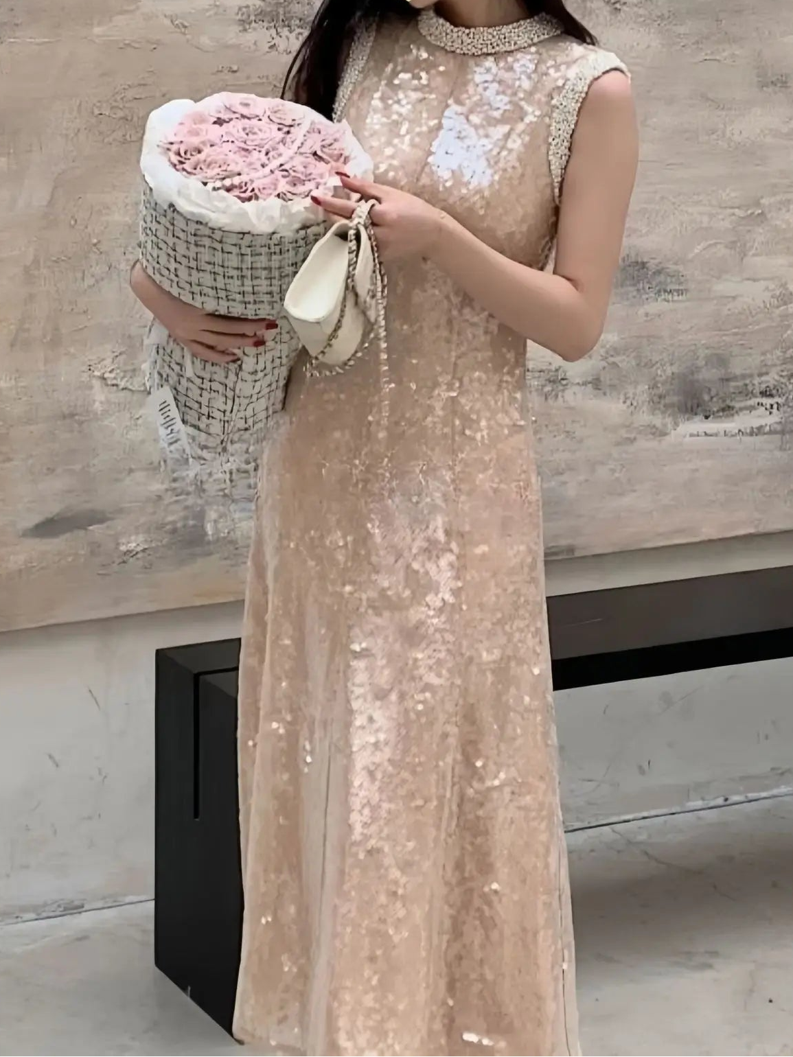 Woman in a beige sequin dress holding a floral arrangement and handbag against a neutral background
