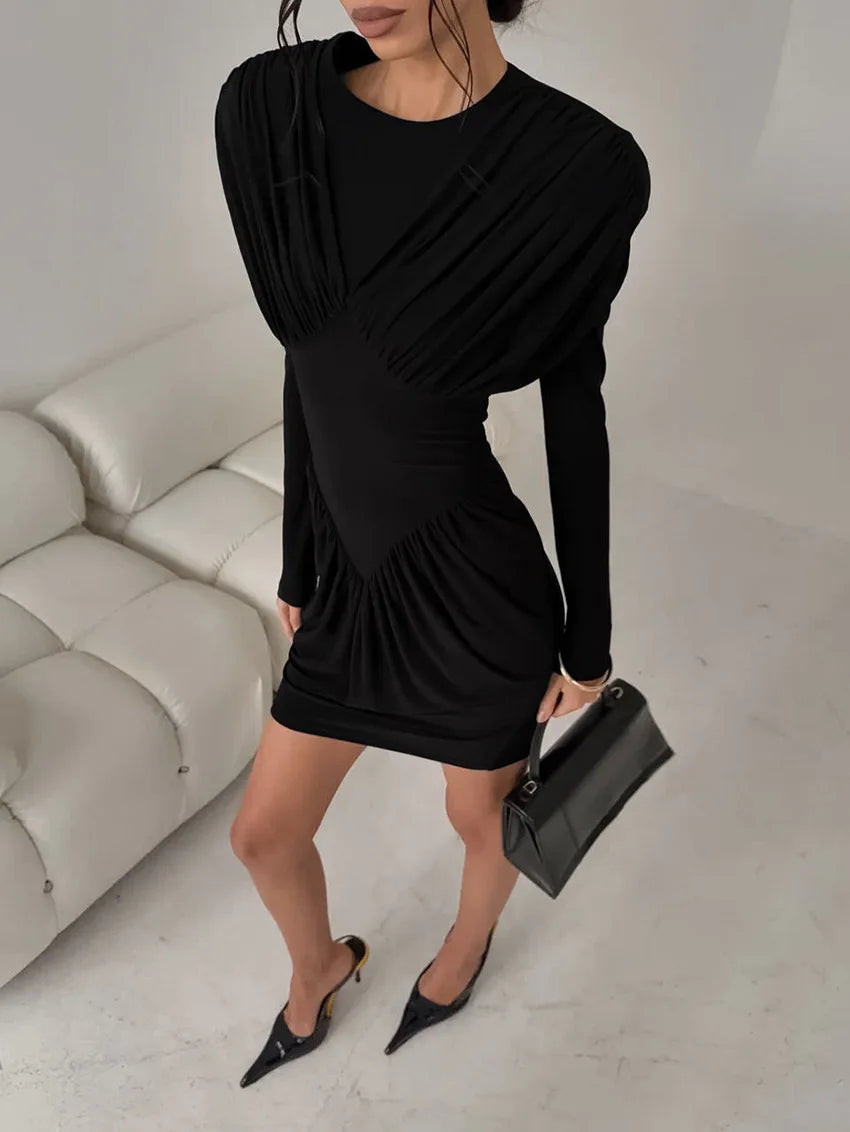 Woman wearing a black dress holding a black handbag in a minimalistic room.
