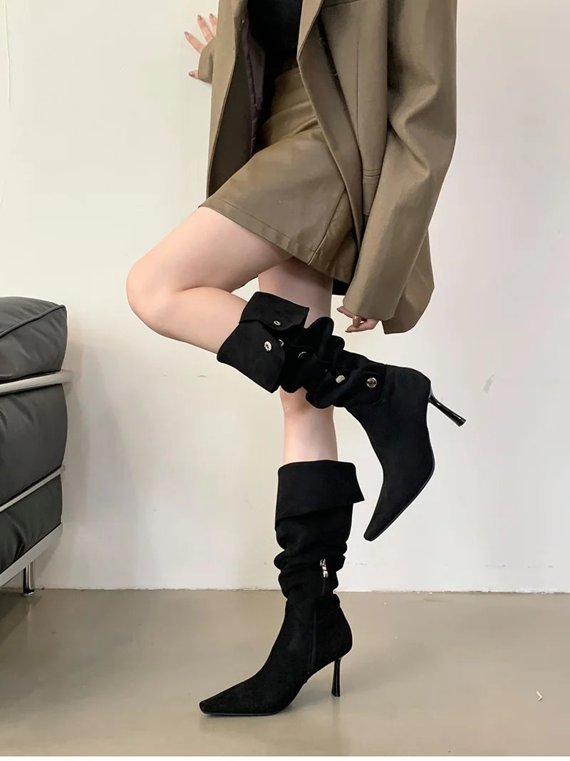 Person wearing black knee-high boots with high heels, standing against a plain background.