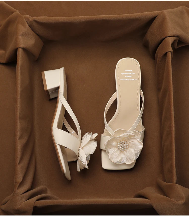White high-heeled sandals with floral decorations on a brown fabric background