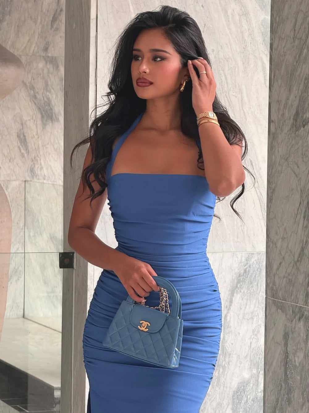 Woman in a blue dress holding a Chanel handbag against a marble wall.