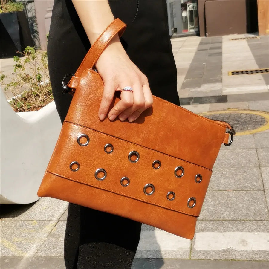 Brown leather clutch bag with grommet details held by a person on a city street.