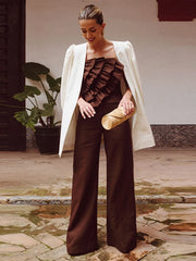 Woman in a brown outfit with ruffled top and wide-leg pants, holding a gold clutch, standing in an outdoor setting.