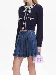 Person wearing a navy blue cardigan with decorative buttons and a pleated denim skirt, holding a pink bag.