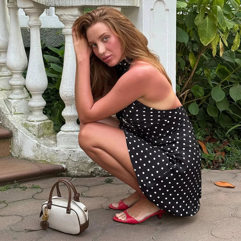 Woman in a black polka dot dress squatting next to a white handbag outdoors.