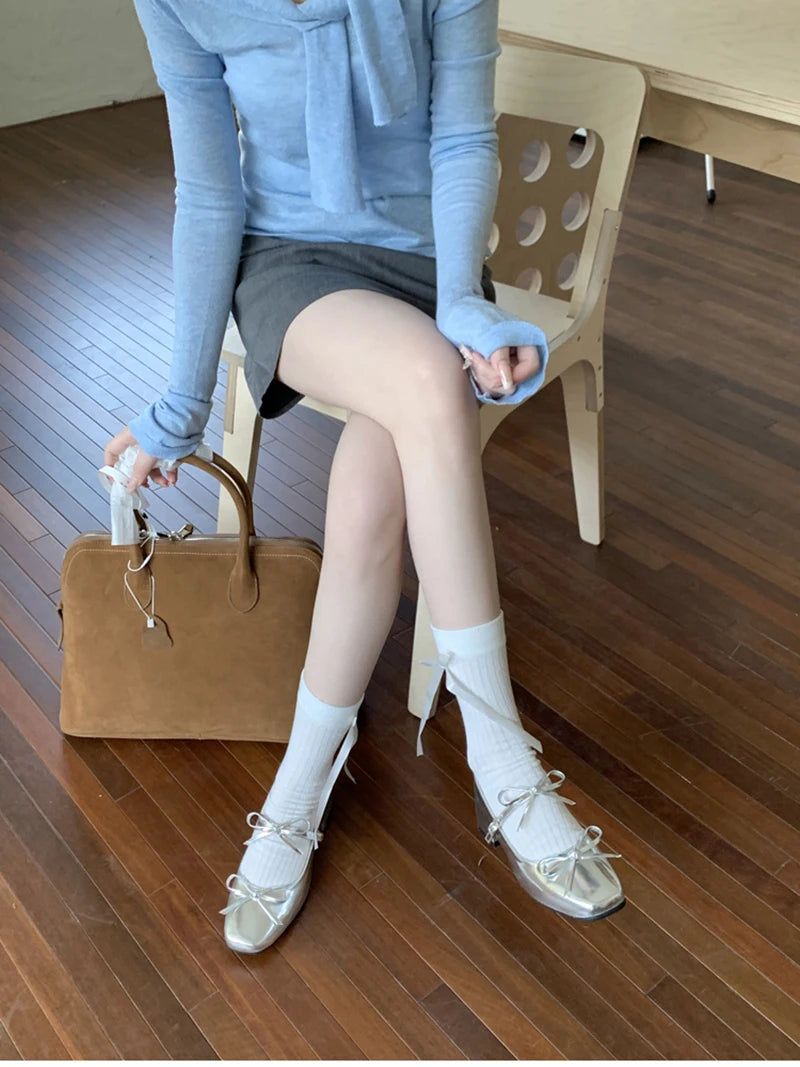Person sitting on a chair wearing a light blue sweater, gray skirt, white socks, and gold shoes with a brown bag beside them.