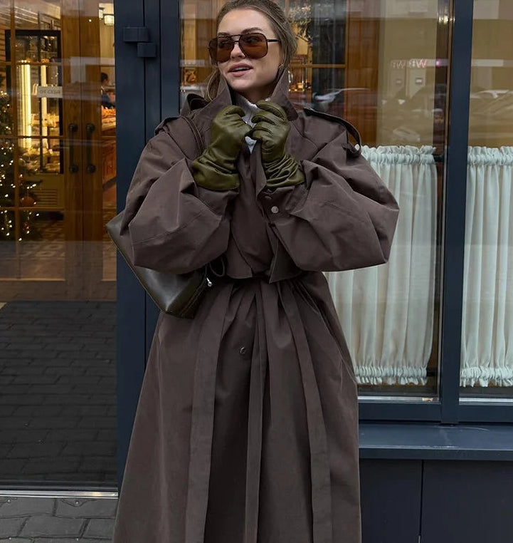 Woman in a long brown coat and sunglasses standing in front of a store window.