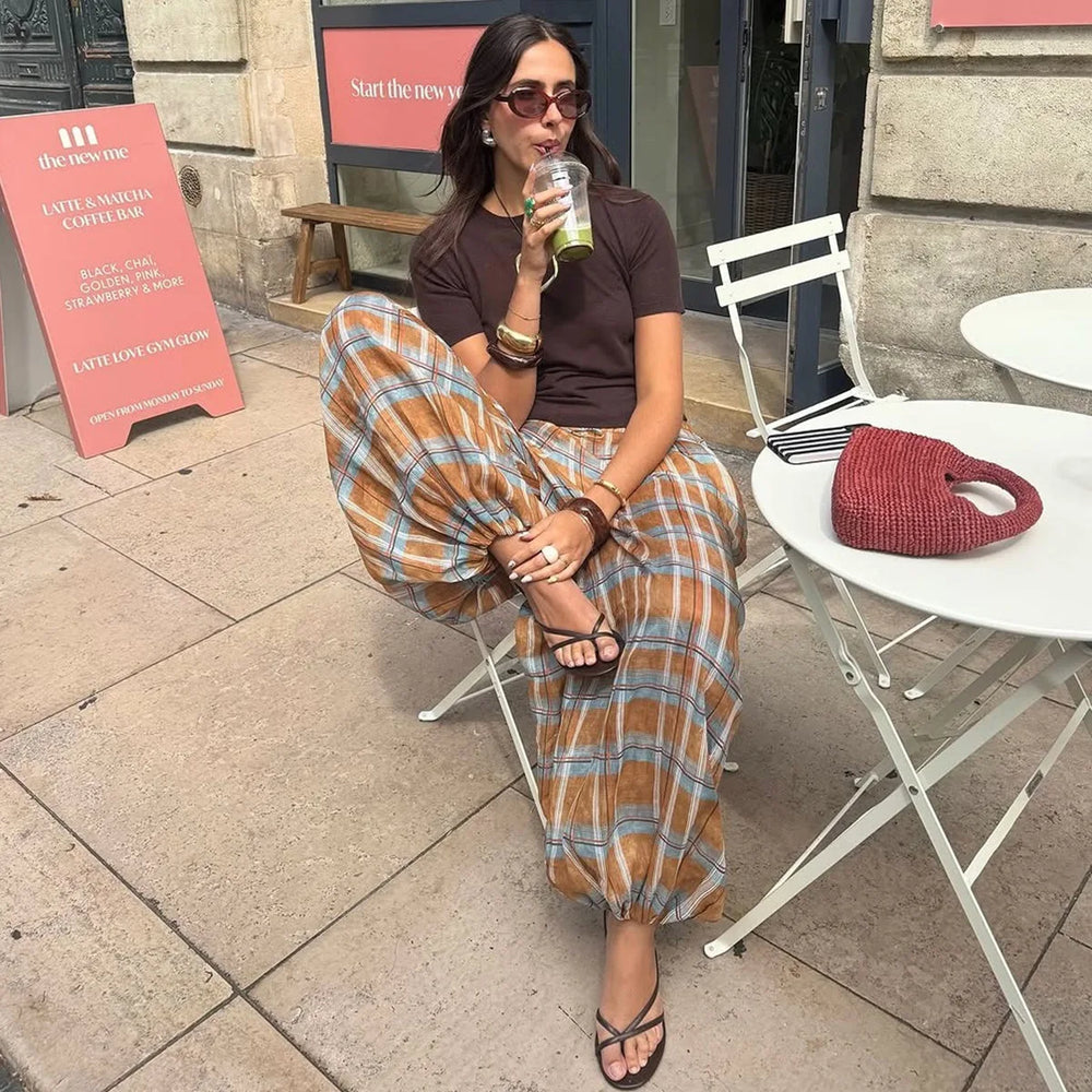 Woman sitting outdoors drinking a beverage, wearing sunglasses and a brown top with plaid pants.