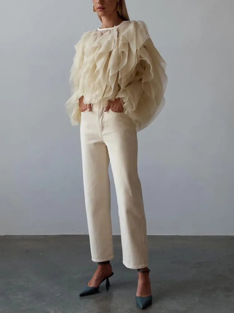 Person wearing a beige ruffled blouse and white pants against a plain background