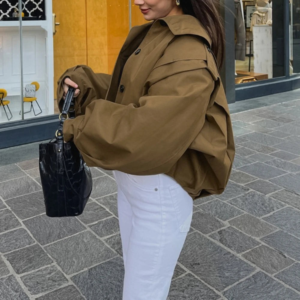 Woman in a brown jacket and white pants holding a black handbag on a city street.