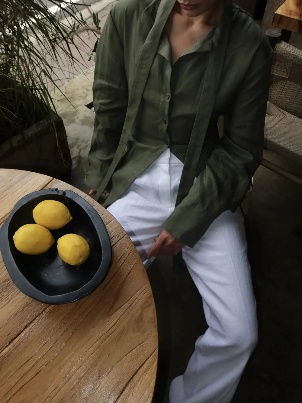 Person in a green jacket and white pants standing next to a table with lemons in a black container.