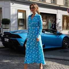 Woman in a blue dress standing in front of a blue sports car on a city street.