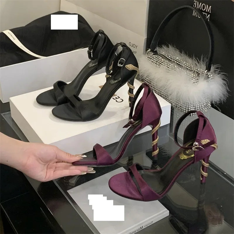 Purple high-heeled shoes with gold accents held by a hand, with black shoes and decorative bag in the background.