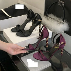 Purple high-heeled shoes with gold accents held by a hand, with black shoes and decorative bag in the background.