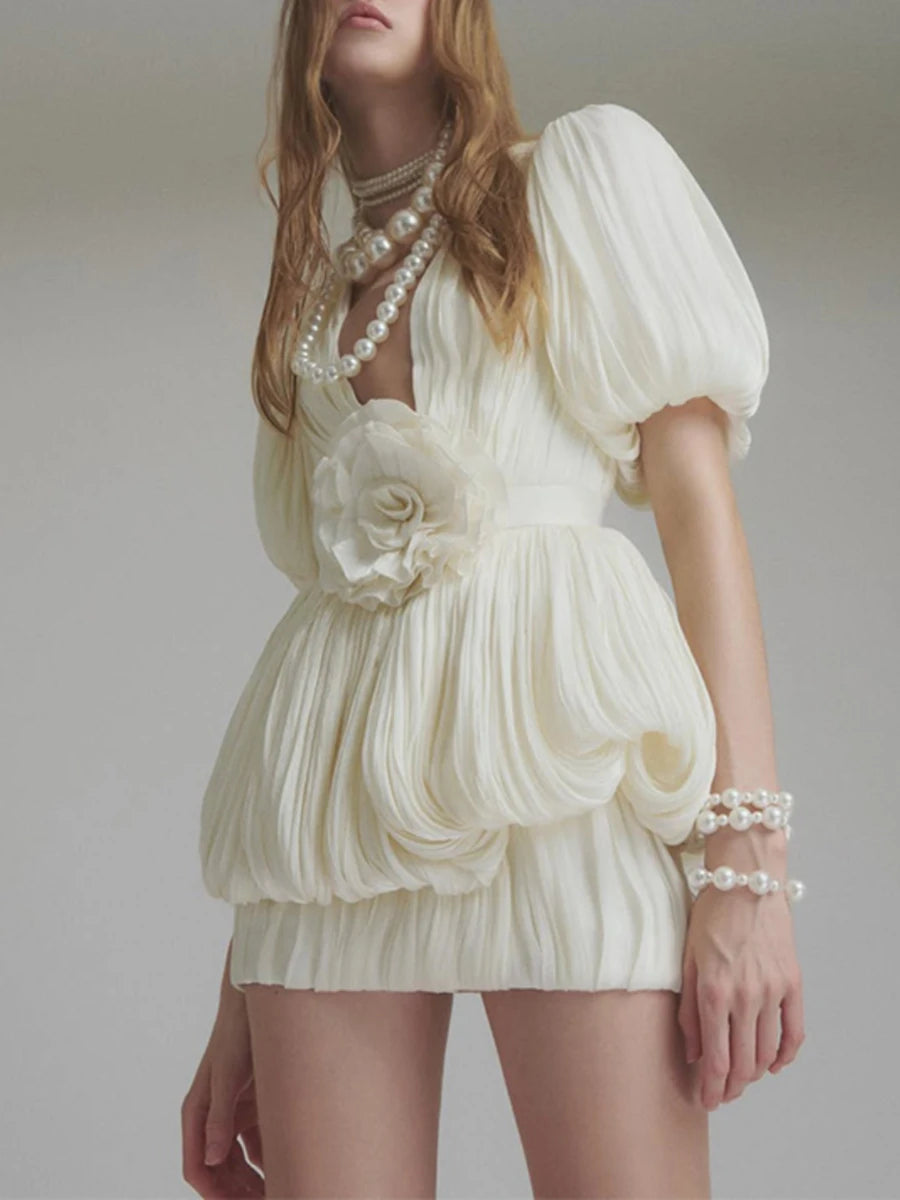 Woman wearing a white dress with floral details and pearl accessories against a plain background