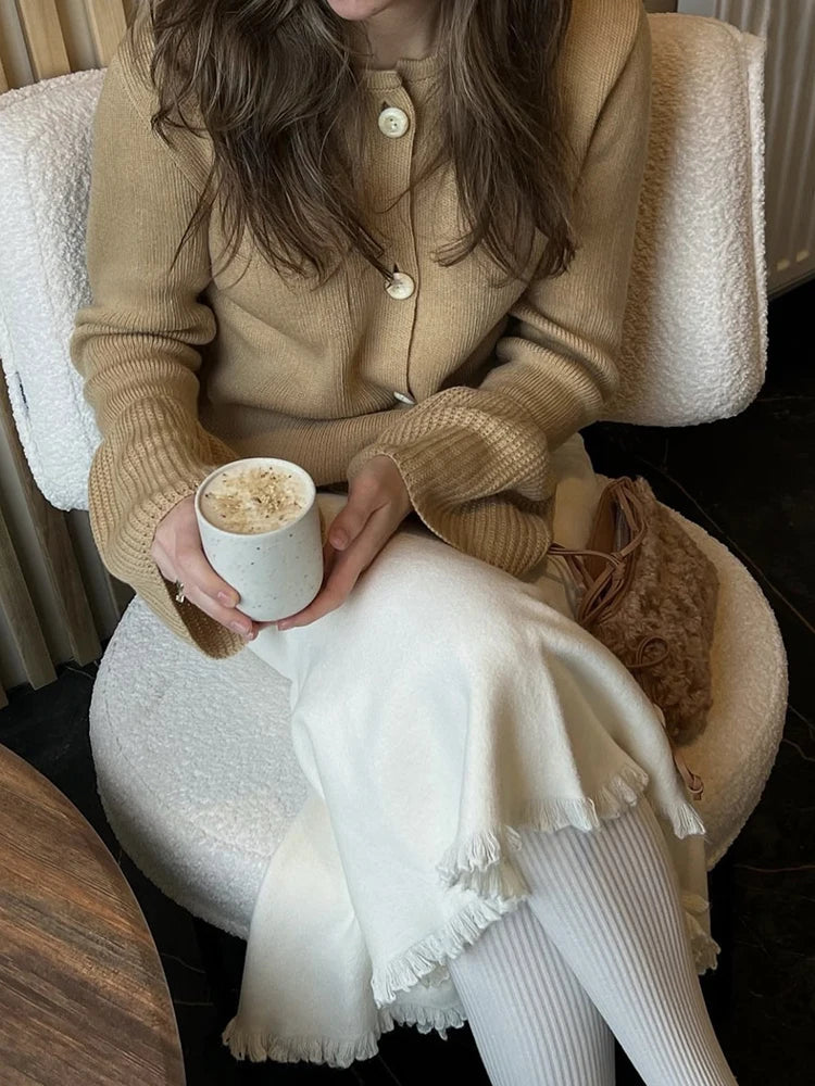 Person wearing a beige coat and white pants, holding a cup, sitting on a chair.