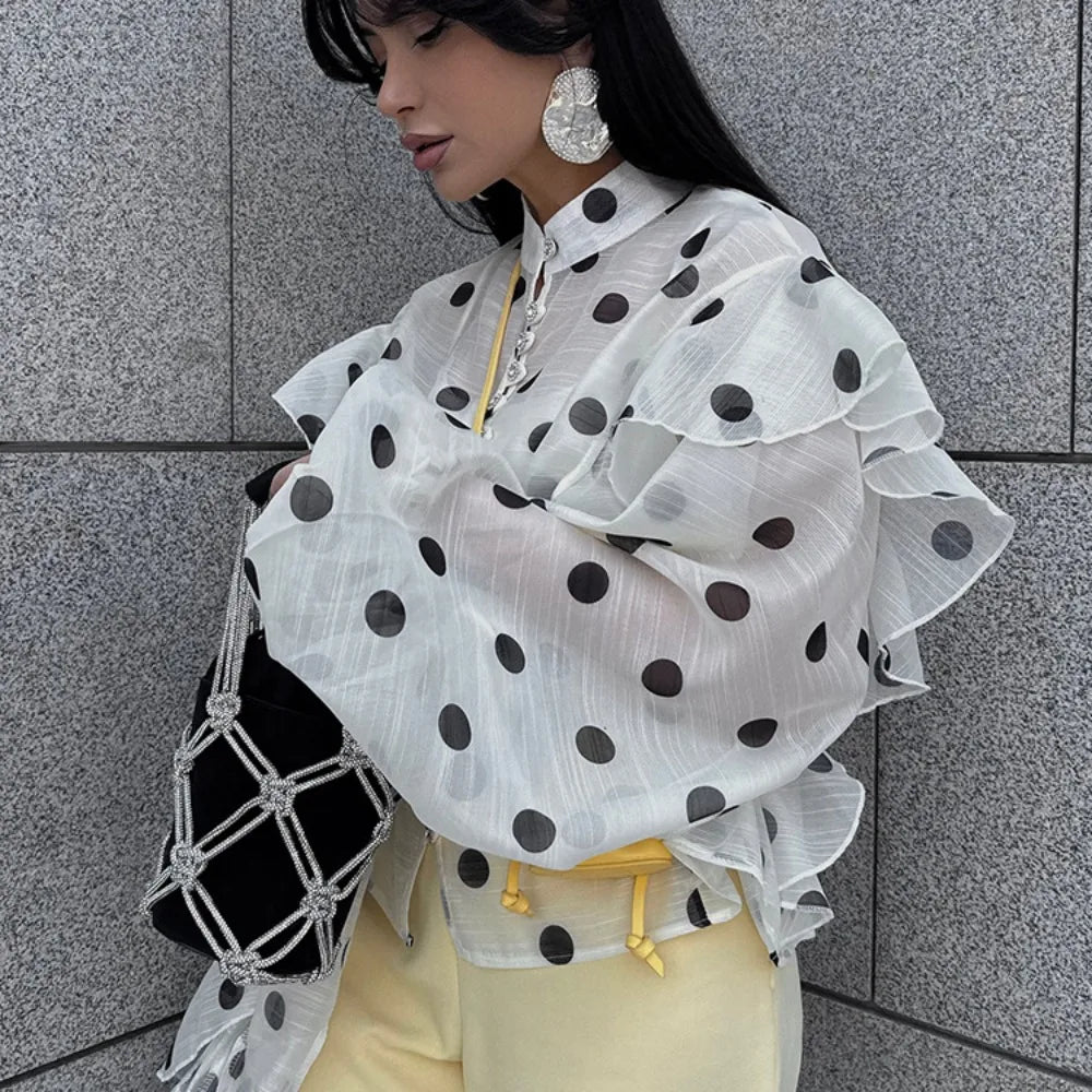 Person wearing a white blouse with black polka dots and yellow pants against a gray tiled wall.