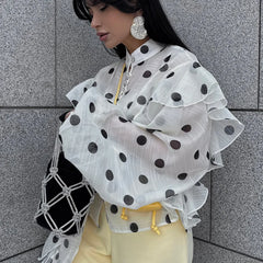 Person wearing a white blouse with black polka dots and yellow pants against a gray tiled wall.