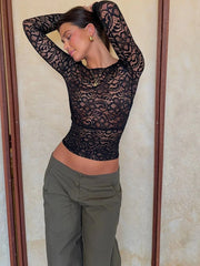 Person wearing a black lace top and green pants against a beige wall.