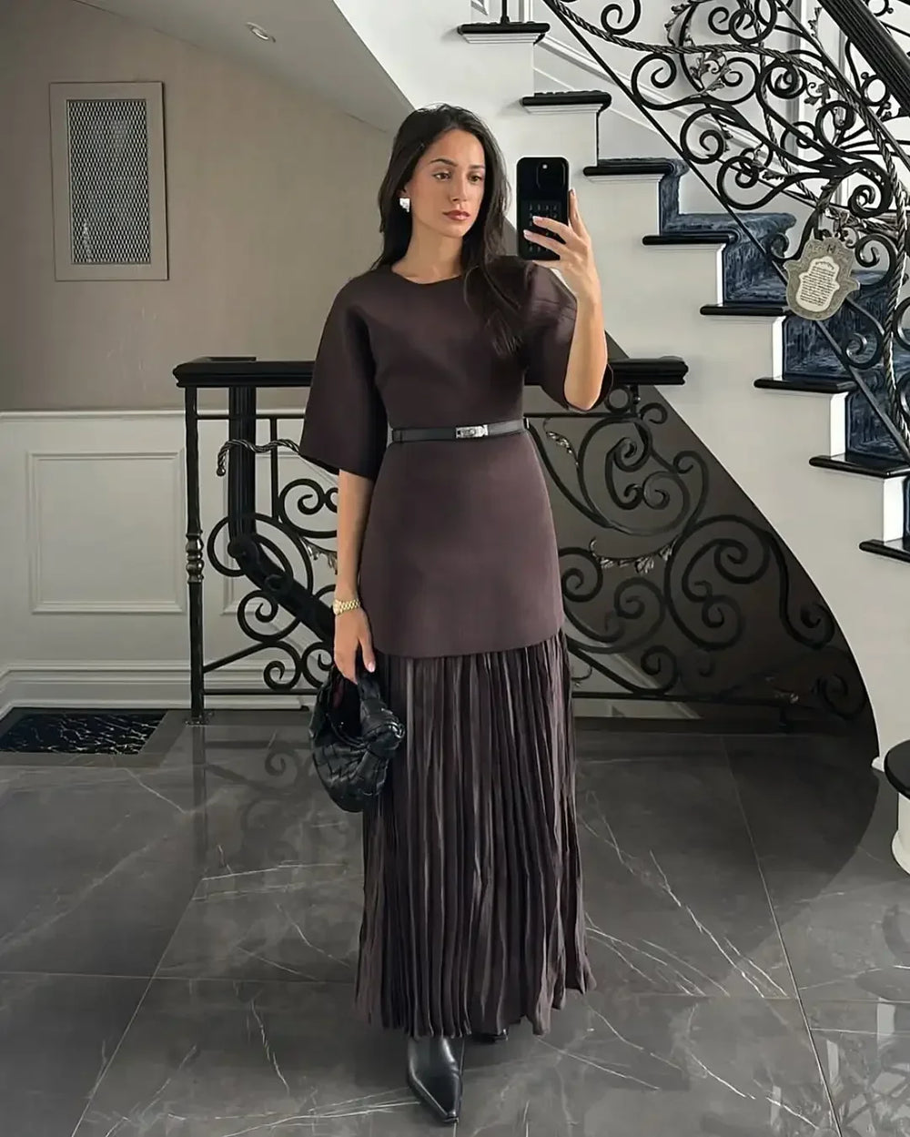 Woman taking a mirror selfie in a dark brown dress with a pleated skirt in a staircase setting.