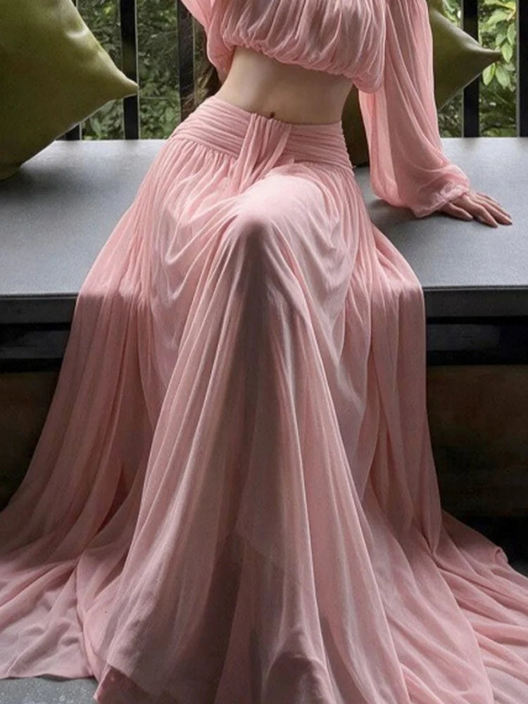 Pink off-shoulder chiffon crop top and maxi skirt with ruched waist detail, woman seated.