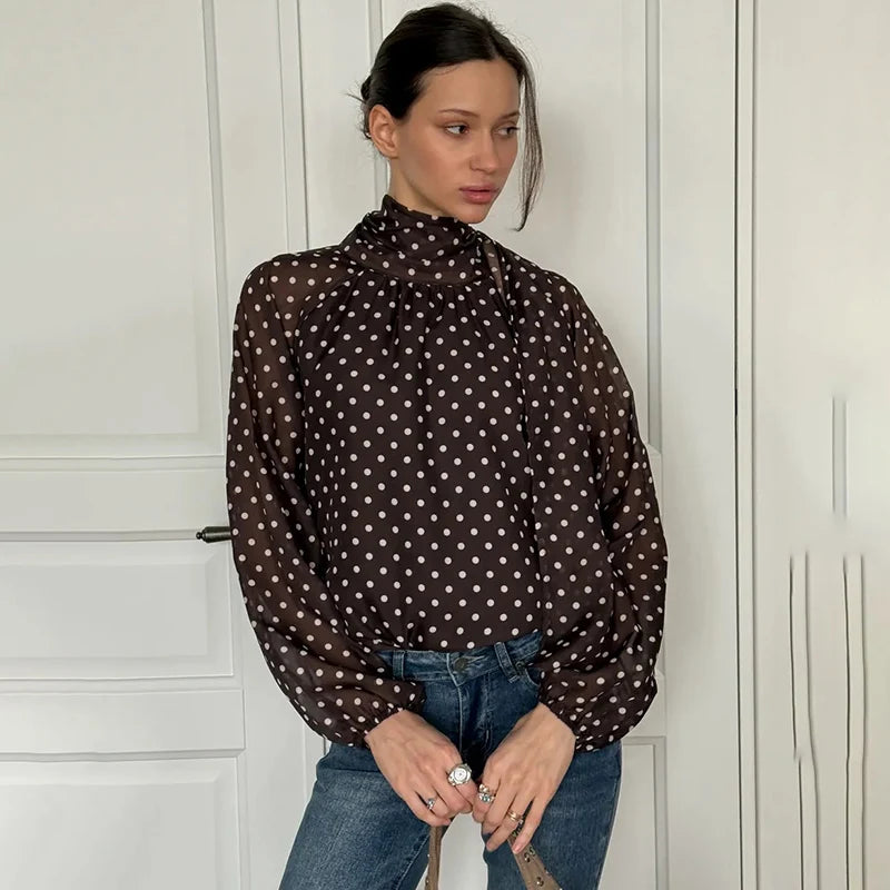 Woman wearing a polka dot blouse and jeans against a white wall.