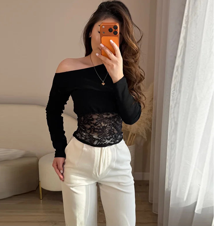 Woman taking a mirror selfie wearing a black off-shoulder top with lace detail and white pants.
