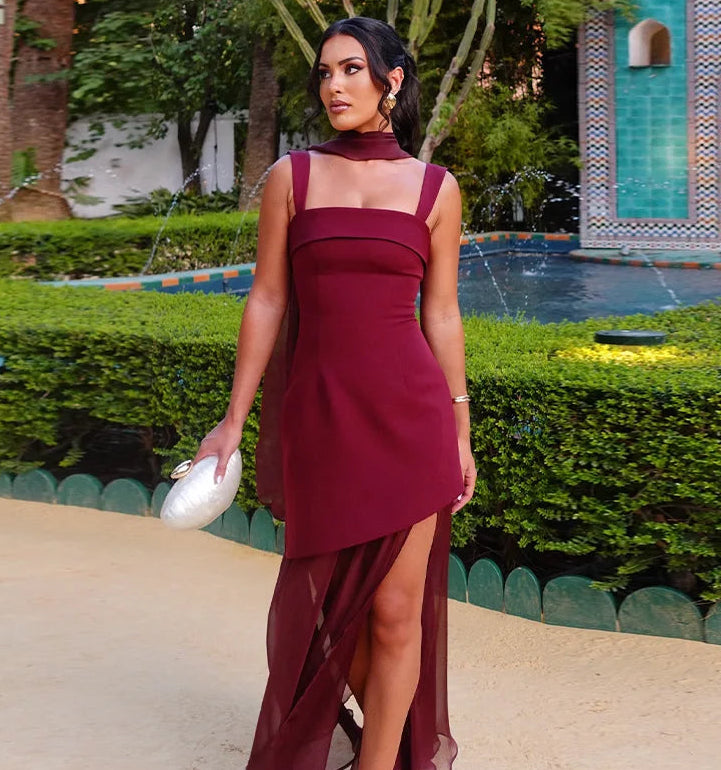 Woman in a burgundy dress standing in an outdoor setting with greenery and a decorative wall.