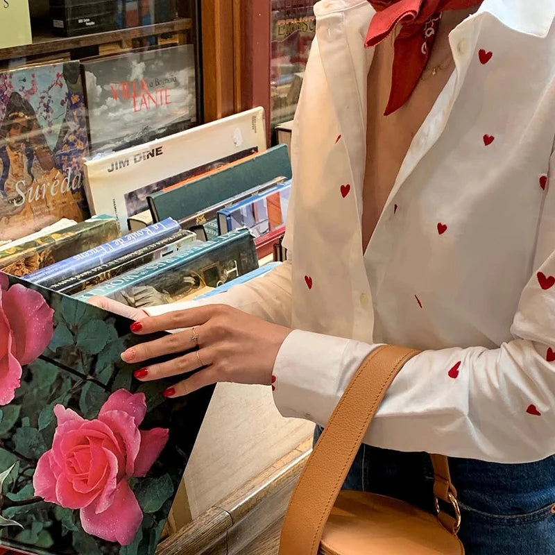 Person wearing a white blouse with red heart patterns, browsing vinyl records in a store.