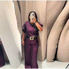 Person wearing a purple outfit with a belt, standing in front of beige curtains.