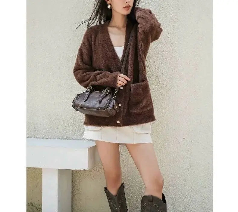 Woman wearing a brown cardigan, white skirt, and black boots holding a black handbag against a light background.