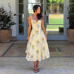 Woman in a floral dress standing in front of a glass door with plants around.
