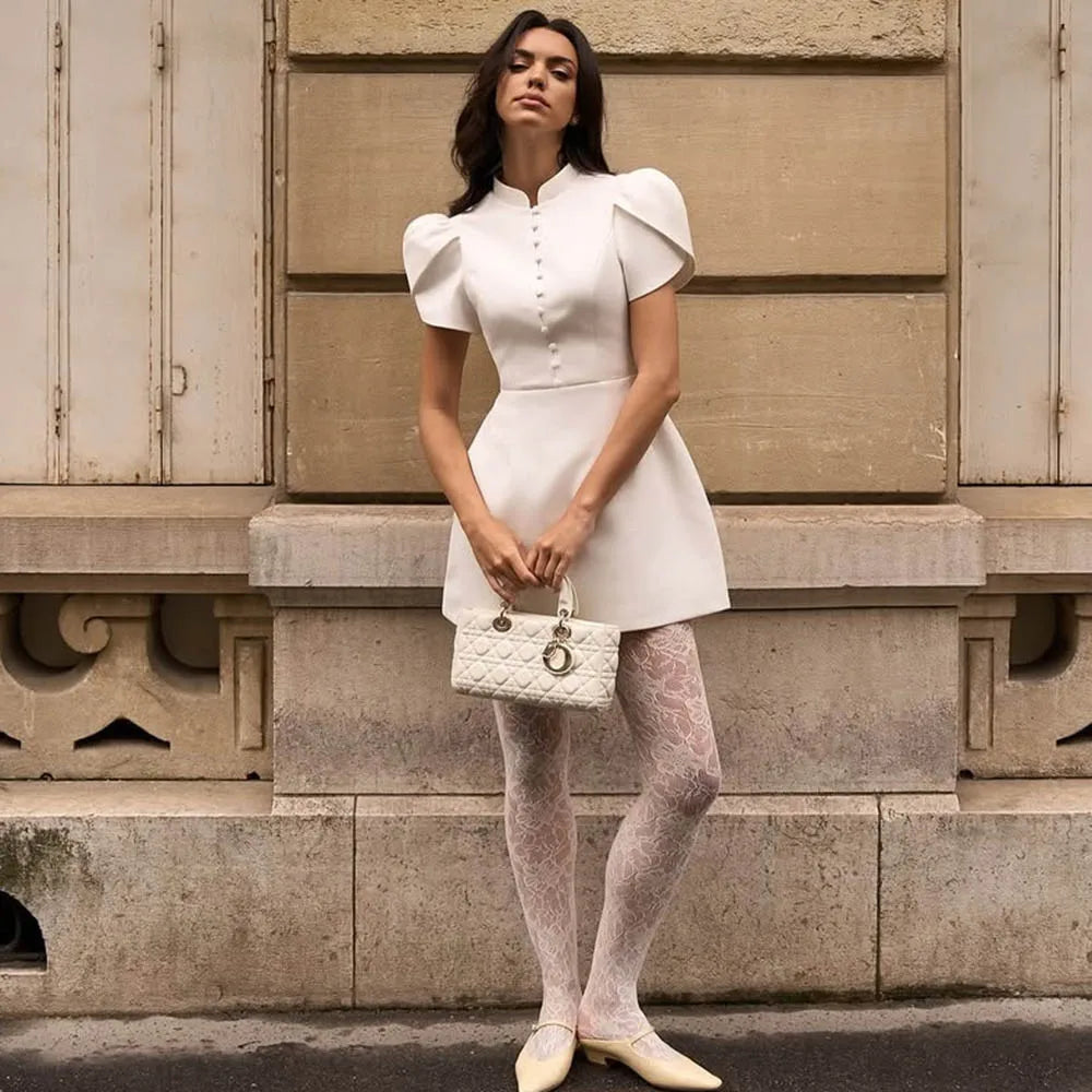 Woman in a white dress holding a handbag against a beige wall.