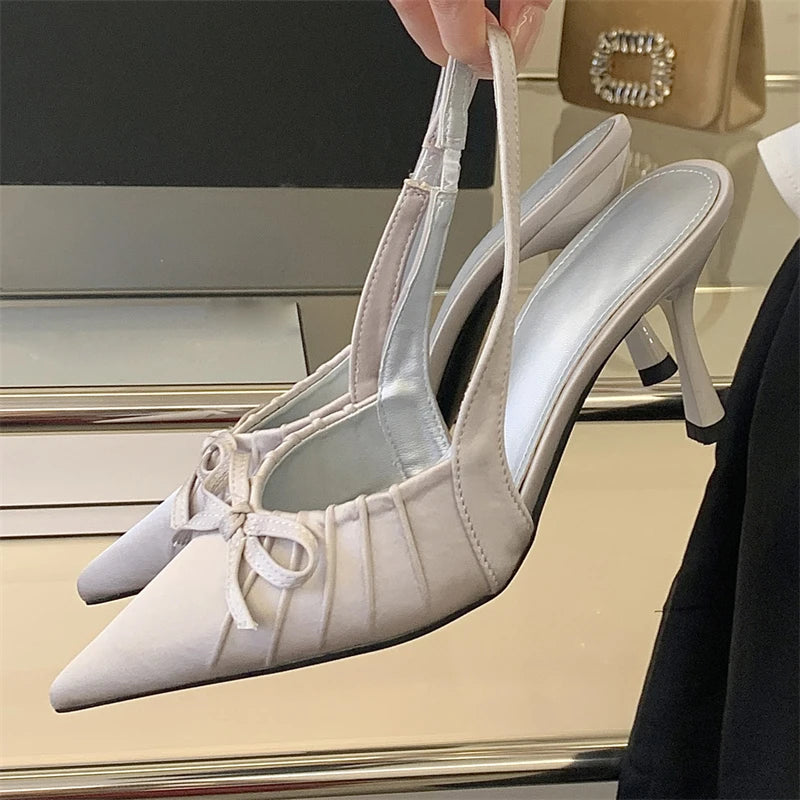 White heeled shoes with ribbon details held by a hand in front of a car.