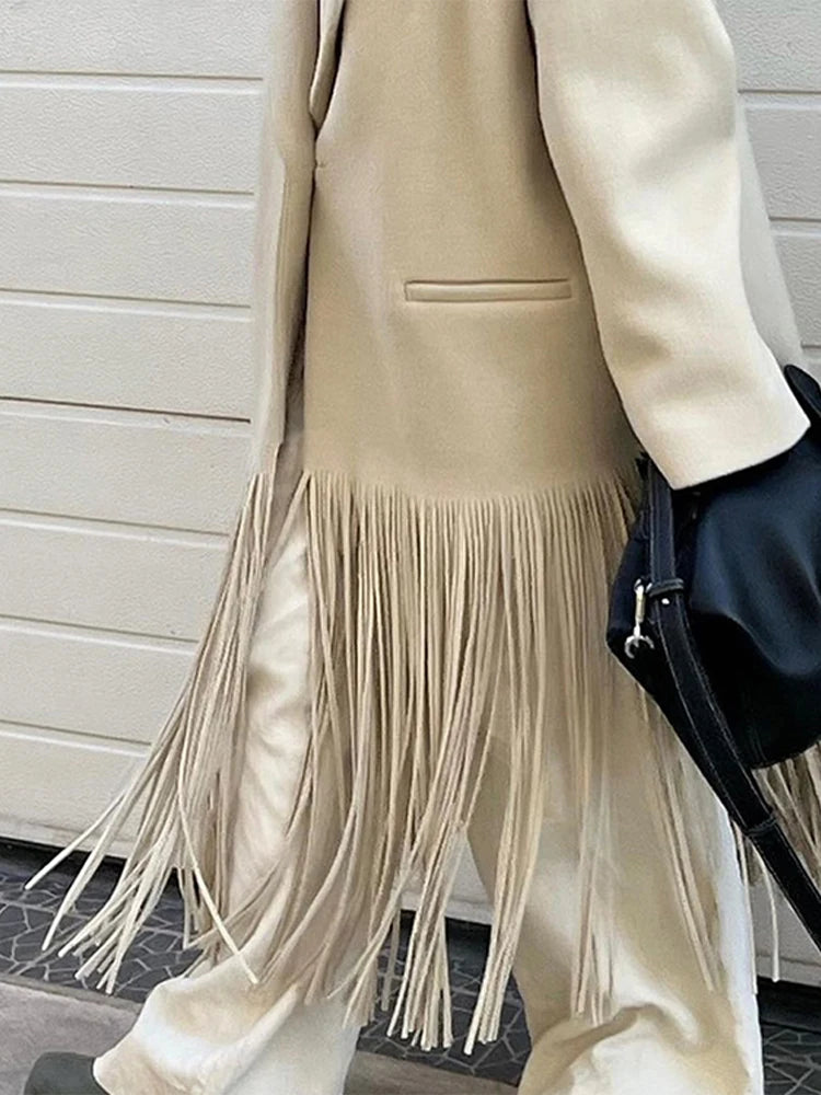 Person wearing a beige coat with fringe details against a light-colored wall.