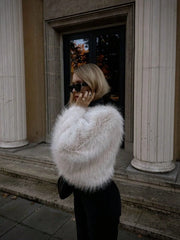 Person wearing a white fur coat standing in front of classical architecture.