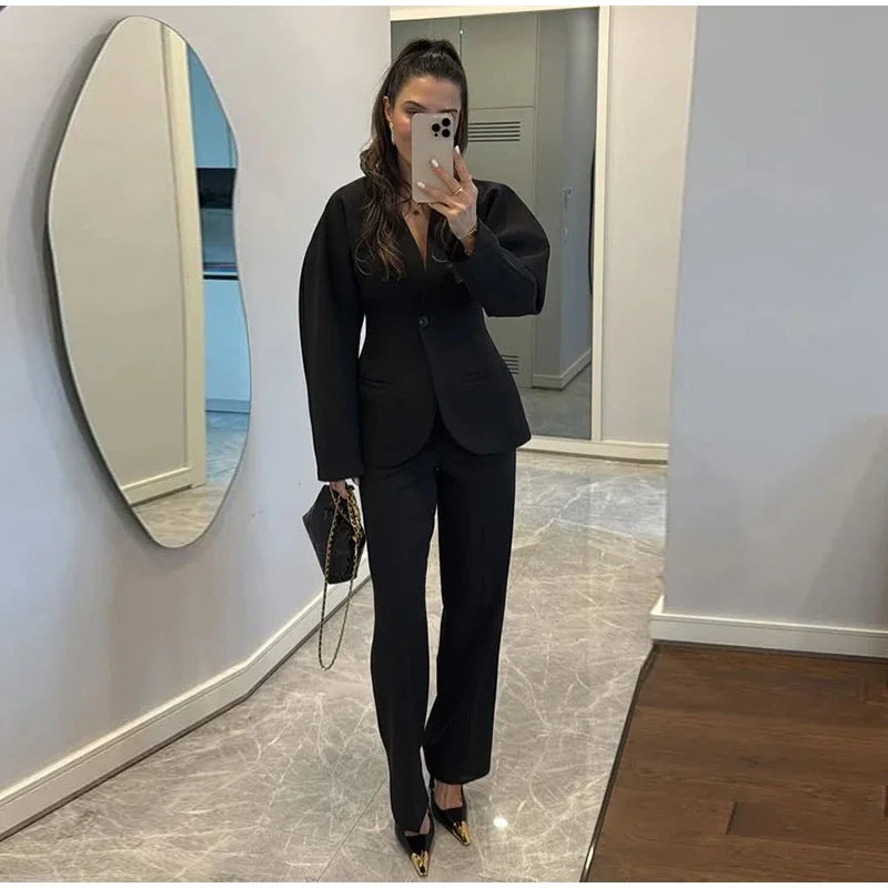 Woman taking a mirror selfie wearing a black suit in a room with a marble floor and white walls.