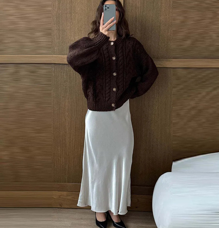 Person wearing a brown cardigan and white skirt against a wooden wall.