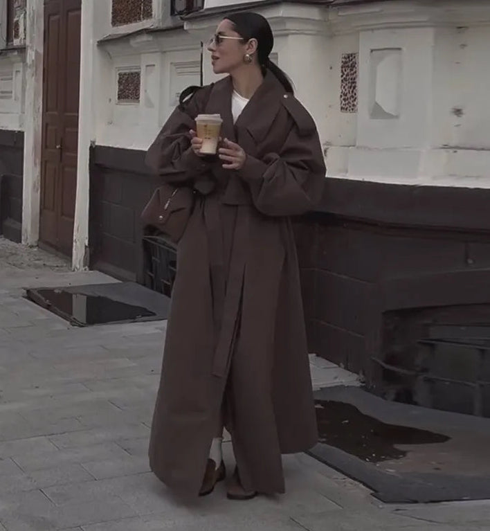Woman in a long brown coat holding a coffee cup on a street.