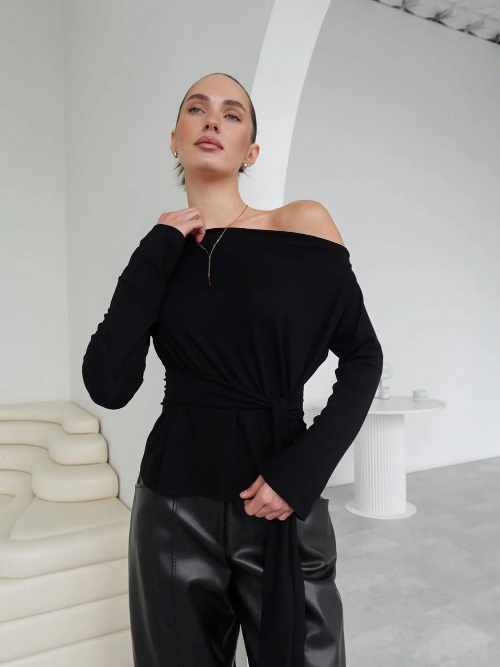 Person wearing a black off-shoulder top and leather pants in a minimalistic setting