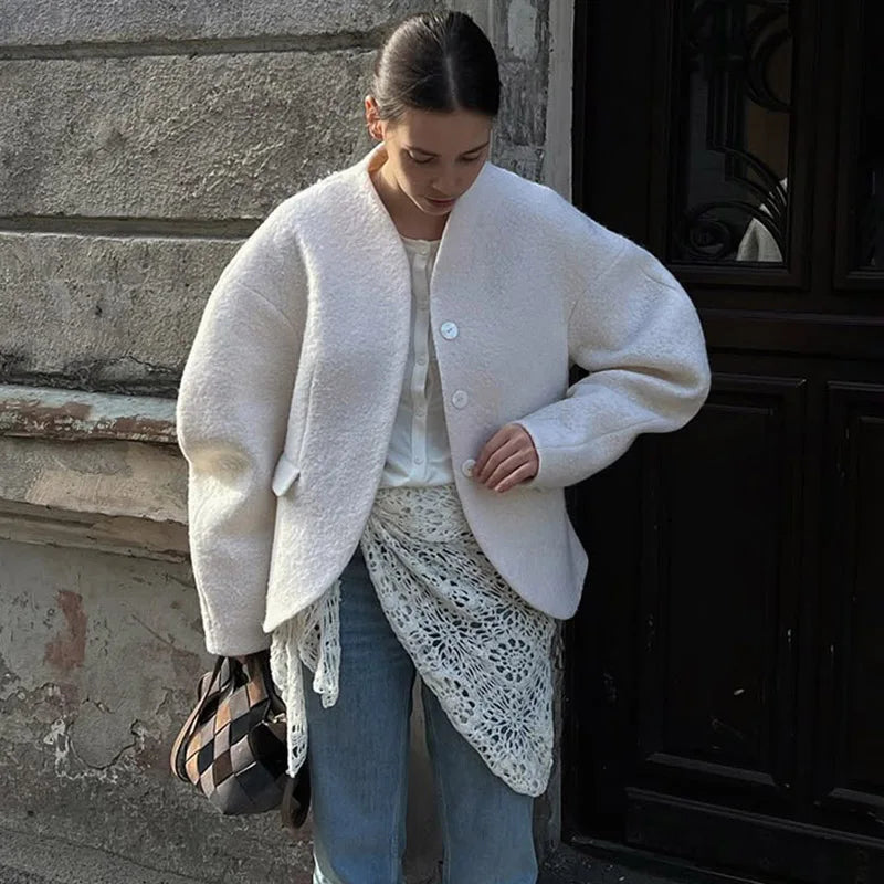 Woman wearing a white coat and blue jeans standing against a textured wall.