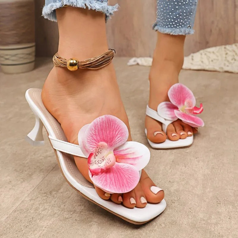 White high-heeled sandals with pink floral decorations worn by a person on a neutral background.
