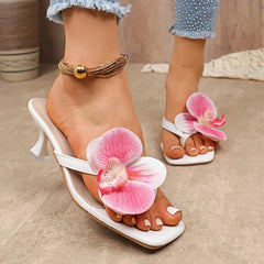 White high-heeled sandals with pink floral decorations worn by a person on a neutral background.