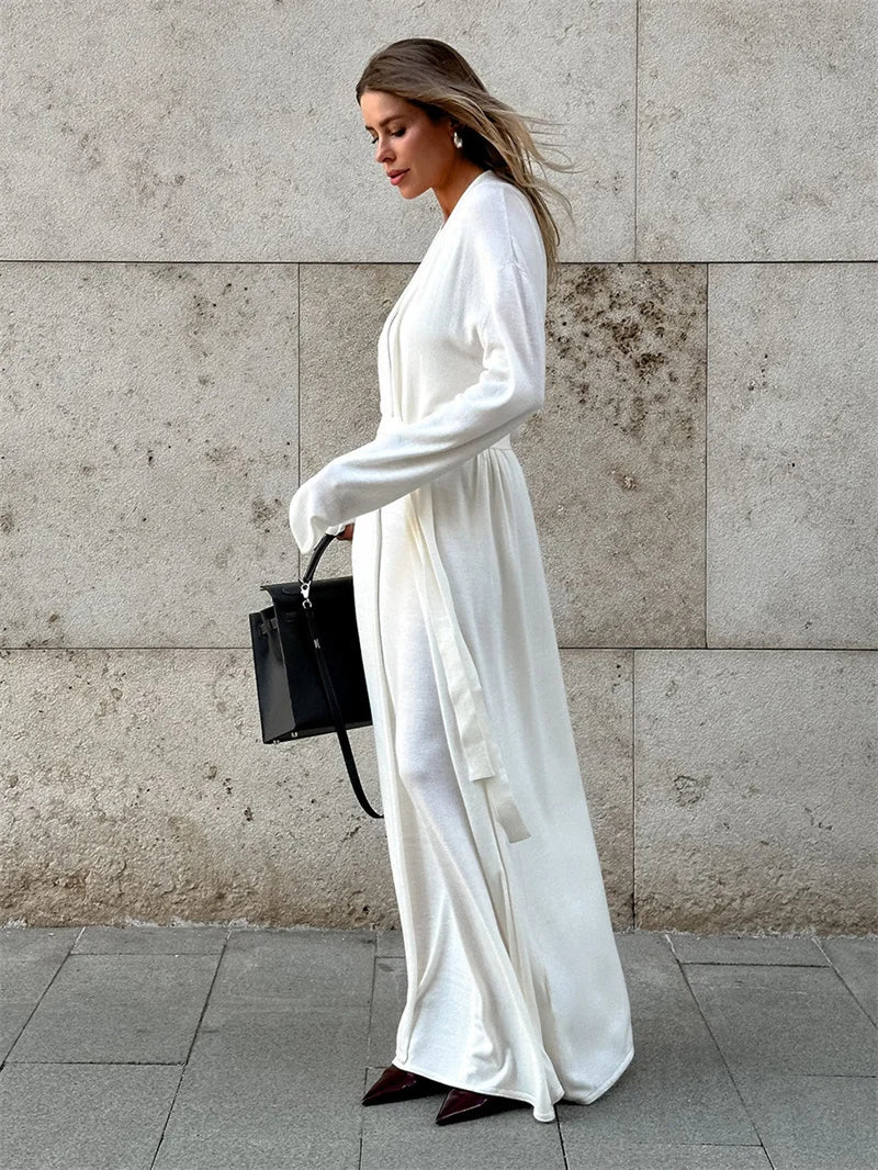 Woman in a long white dress holding a black handbag against a gray wall.