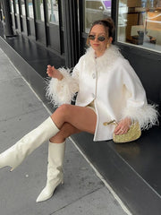 Woman in a white coat with feather trim and white knee-high boots sitting on a bench.