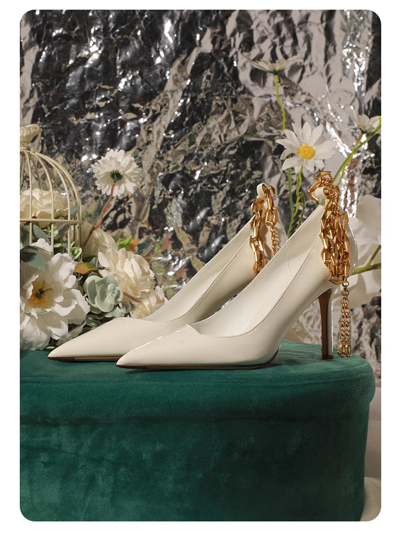 White high-heeled shoes with gold chain details on a green surface with floral and metallic background