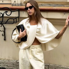 Woman in a stylish outfit with a white top and pants, holding a black clutch against a neutral wall.
